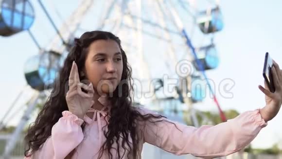 一个穿着粉红色连衣裙留着长发的年轻女孩站在摩天轮附近用手机自拍慢动作视频的预览图