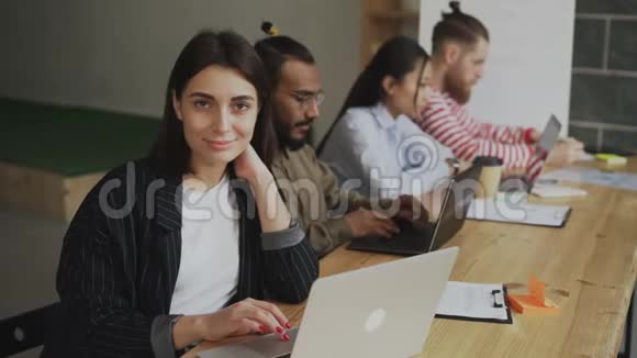 微笑的女性自由职业者看着坐在繁忙的办公室里的手提电脑相机的画像多重视频的预览图
