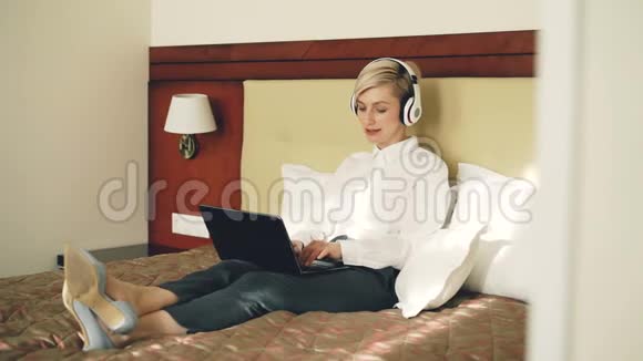 快乐的女商人戴着耳机在笔记本电脑上工作一边听音乐一边笑着跳舞一边躺在床上视频的预览图