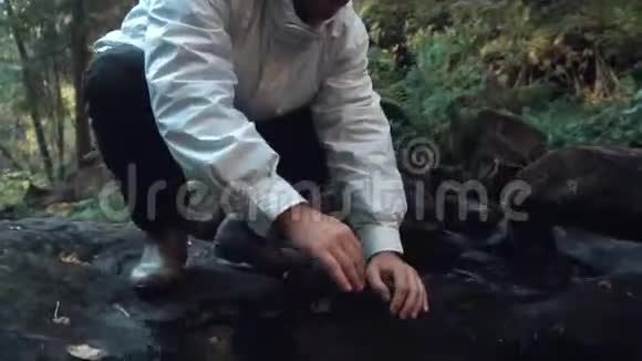穿着白色夹克的人在秋天的森林里喝着小溪里的淡水录像特写视频的预览图
