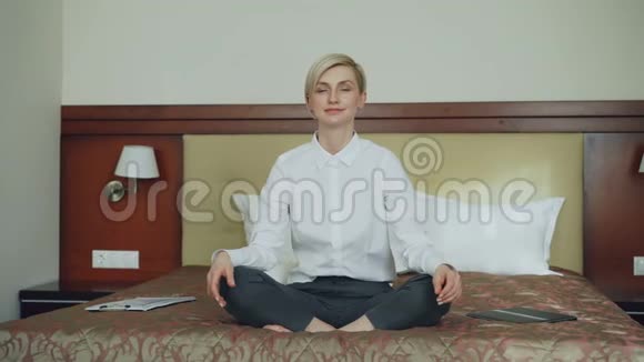 微笑的女商人坐在酒店房间的床上放松瑜伽莲花的位置旅游商务和人员视频的预览图