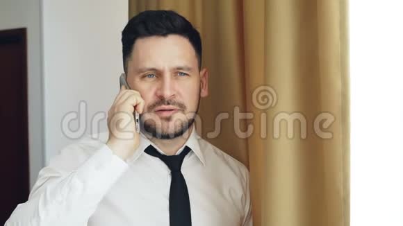 穿着白色衬衫和领带的成年胡子商人站在酒店房间的窗户附近用手机交谈视频的预览图