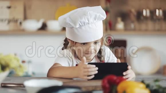 可爱的小女孩在早餐时使用智能手机坐在厨房里她穿着围裙看着视频的预览图