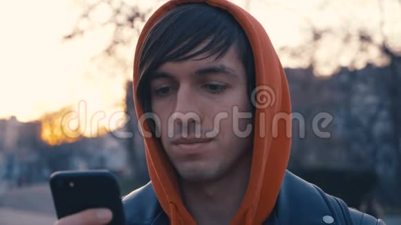 英俊的年轻人在阳光城市街道城市背景穿红色连帽衫的人用智能手机日落背景视频的预览图