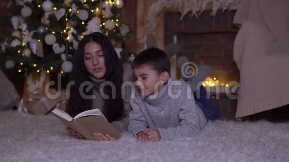 在圣诞节留着长发的妈妈给儿子读一本书亲吻他躺在圣诞树旁的地板上视频的预览图