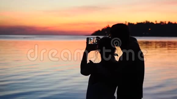 海滩上年轻夫妇的剪影在日落时拍了一幅自拍肖像慢动作享受度假理念的人视频的预览图