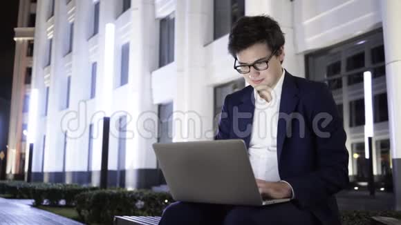 一个年轻的商人晚上在市中心的长椅上用笔记本电脑工作视频的预览图