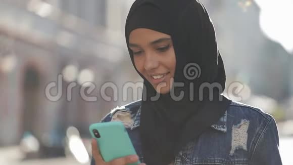 年轻微笑的穆斯林妇女戴着头巾站在市中心使用智能手机通信视频的预览图