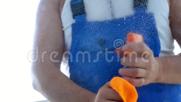 特写镜头透过窗户观看男清洁服务工人穿蓝色工作服喷涂清洁窗口视频的预览图