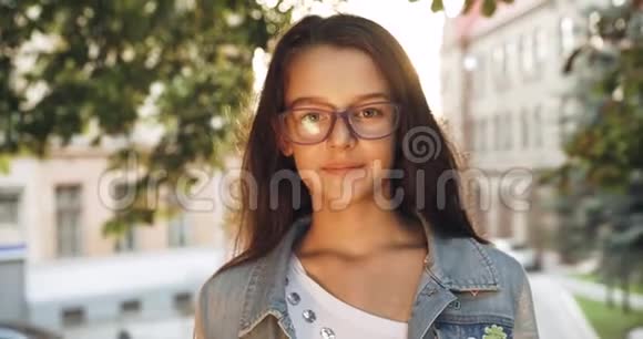 带着紫色眼镜的深色长发的积极的小女孩正对着镜头微笑城市背景太阳耀斑4k视频的预览图