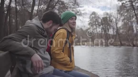 一对年轻的游客背着背包坐在河岸的春天公园的长凳上有胡子的人视频的预览图
