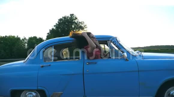 迷人的女孩戴着太阳镜斜靠在老式车窗外享受旅行年轻夫妇乘坐复古车旅行视频的预览图