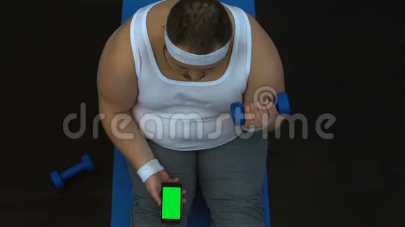 肥胖男子举起哑铃观看健身视频智能手机绿色屏幕视频的预览图