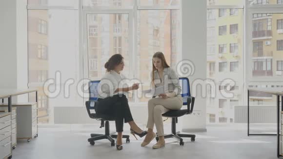 一名妇女解释说办公室工作人员坐着两名年轻妇女坐在办公室椅子上谈判公司的文件视频的预览图