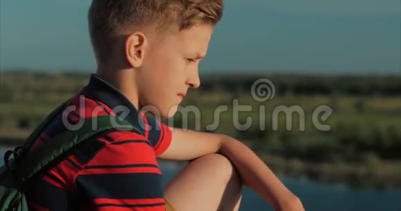 特写肖像白人少年穿着红色衬衫背着背包日落时分坐在一座高山上视频的预览图