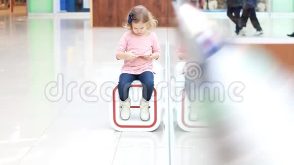 在购物中心坐着玩智能手机的女孩子宝宝在等父母去购物视频的预览图