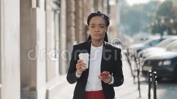 一位穿着西装在城市里走来走去喝咖啡和使用咖啡的严肃的非裔美国女商人的肖像视频的预览图