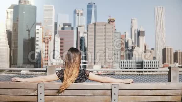 在纽约一位留着长发的快乐女孩坐在长凳上欣赏美丽的曼哈顿天际线风景视频的预览图