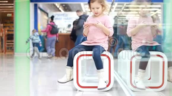 在购物中心坐着玩智能手机的女孩子宝宝在等父母去购物视频的预览图