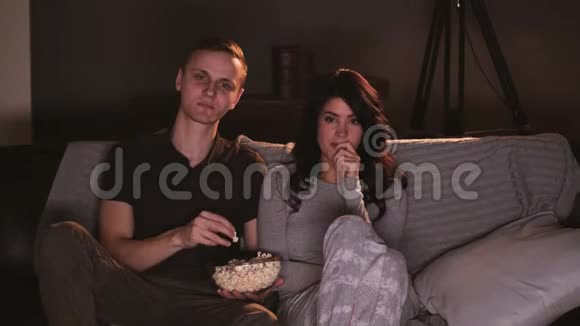 年轻夫妇在家看电视上的滑稽节目吃爆米花视频的预览图