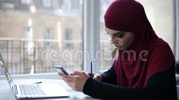 年轻的穆斯林女孩头上戴着紫色头巾坐在灯光下从智能手机上写下一些东西视频的预览图