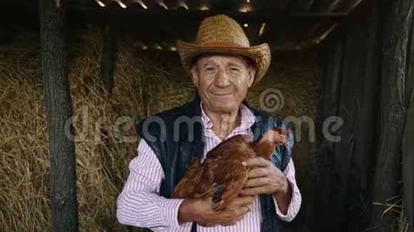 一个戴草帽的老农正在抱一只活鸡干草背景下一个带鸡的人的肖像视频的预览图