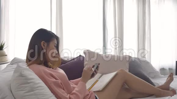 快乐的年轻亚洲女人在家学习在手机上看短信或短信微笑着躺在家里的沙发上视频的预览图
