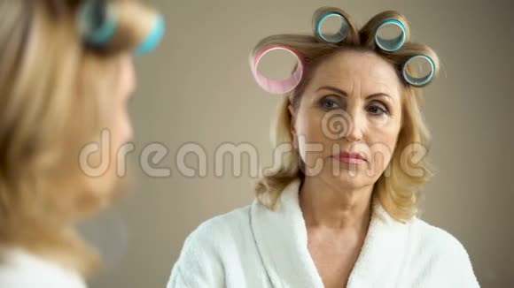 一个年老的悲伤的女人带着卷发和化妆看着镜子的倒影视频的预览图