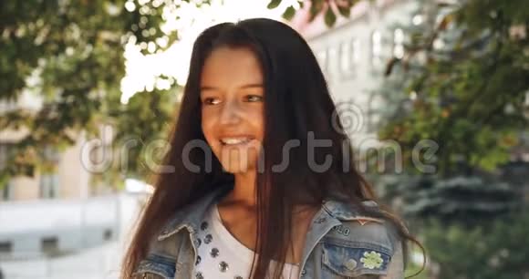 可爱的微笑的小女孩的肖像积极地摇动着她长长的黑发城市景观背景4K录像太阳视频的预览图