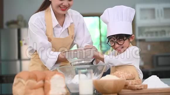 可爱的亚洲小男孩和漂亮的妈妈在家厨房的桌子上用筛子筛玉米粉准备视频的预览图