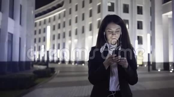 年轻的女商人带着智能手机和耳机走在城市的一条夜晚街道上视频的预览图