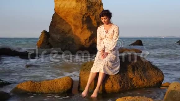 日出时可爱的白种人深褐色的女孩穿着淡粉色的裙子坐在大海的岩石上视频的预览图
