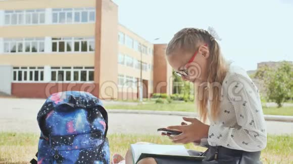 一个11岁的女学生拿着一本书坐在草地上看着电话视频的预览图