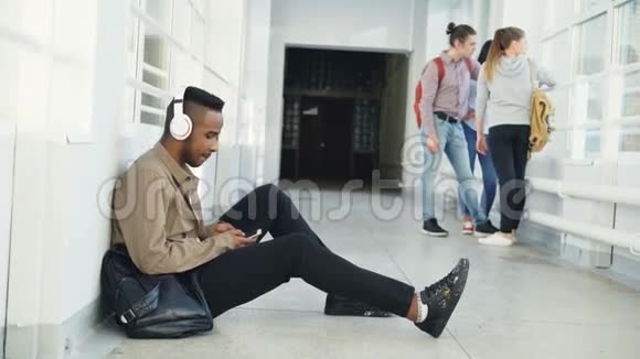 年轻英俊的非裔美国学生坐在白色走廊的地板上头上戴着耳机听音乐视频的预览图