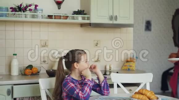 年轻的母亲带着美味的松饼给她可爱的女儿她坐在厨房的桌子旁在家等早餐视频的预览图