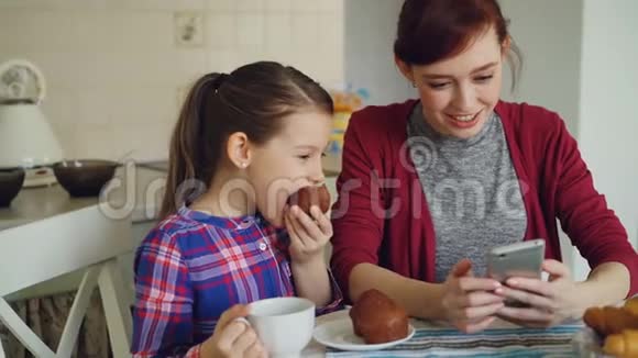 快乐的母亲和可爱的女儿一起浏览智能手机早上在厨房里微笑视频的预览图