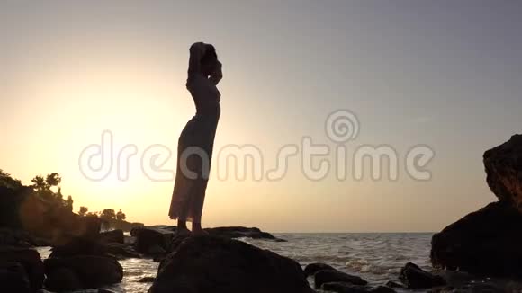 可爱的白种人布鲁内特女孩穿着浅色衣服在日出时在海上的岩石上跳舞视频的预览图
