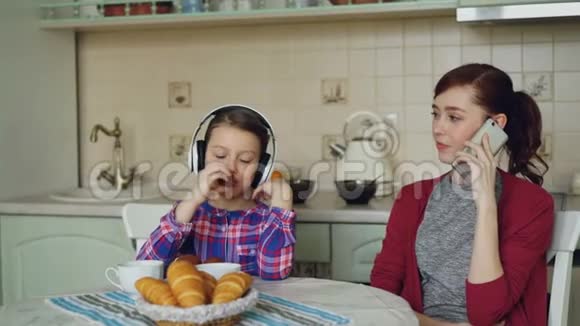 年轻的母亲和可爱的女儿早上在家厨房吃早餐小女孩听音乐视频的预览图