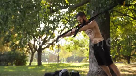 有胡子的年轻肌肉男开始在阳光公园里用健身环进行俯卧撑锻炼4K慢节奏的电影视频的预览图