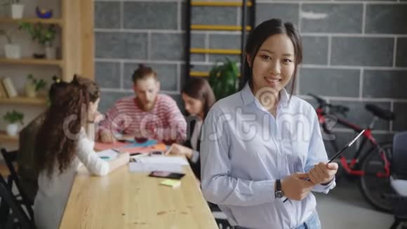年轻的亚洲女企业家手持数码平板电脑在繁忙的创业办公室里看着相机微笑着的画像视频的预览图