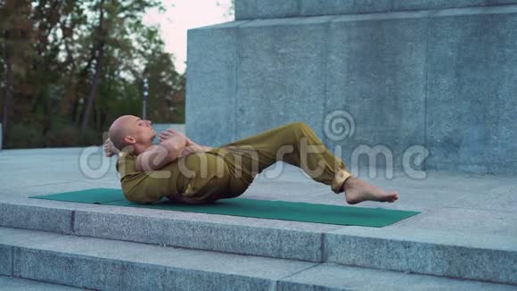 秃头年轻人在城市公园里躺在绿毯上做复杂的瑜伽练习视频的预览图