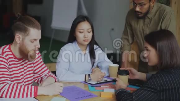 年轻的时髦男人指着一张纸上面写着区块链系统实现计划与坐在旁边的同事分享视频的预览图