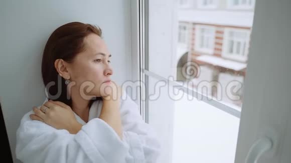 一个穿着白色外套的沮丧的女人坐在窗台上向外看着街道一个悲伤的女人坐在视频的预览图