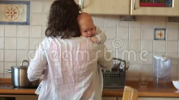 年轻的家庭主妇在厨房准备晚餐一个女人手上抱着一个婴儿多任务她喂养孩子视频的预览图