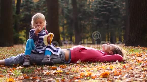 美丽的妈妈和可爱的孩子躺在秋天公园的草地上女儿坐在妈妈身上吃香蕉妈妈望着天空视频的预览图