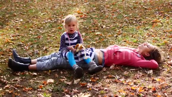 美丽的妈妈和可爱的孩子躺在秋天公园的草地上女儿坐在妈妈身上吃香蕉妈妈望着天空视频的预览图