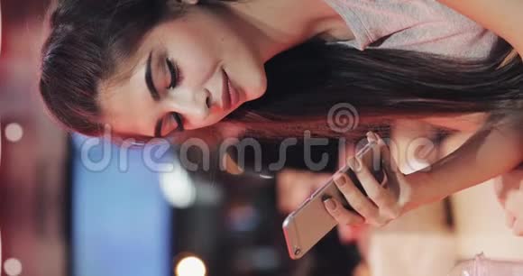 垂直视频年轻迷人的女人美丽的微笑是使用智能手机在晚上坐在酒吧里的女孩视频的预览图