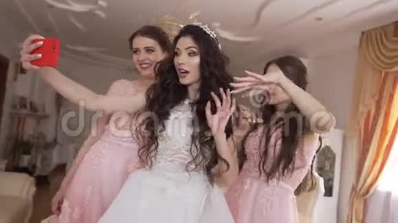 漂亮的年轻新娘有着漂亮的妆容和长长的卷发在家里和两个可爱的伴娘一起玩滑稽的自拍视频的预览图