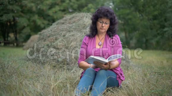 一位老年妇女在干草堆背景下的森林里看一本坐在草地上的书视频的预览图