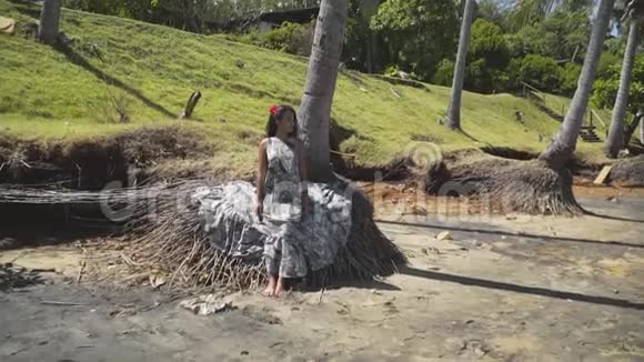 亚洲女孩坐在棕榈树的根部穿着灰色的衣服摆姿势视频的预览图